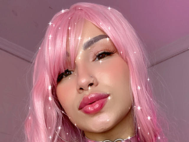 OnniOconor with pink wig, nose ring, and glittery face, pouty pink lips, dark eyebrows, and long eyelashes, looking down, sparkling effects.