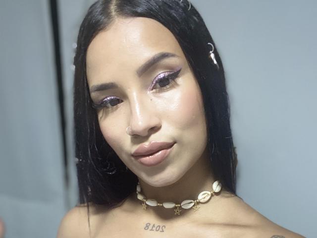 OnniOconor with shiny purple eyeshadow, nose ring, and shell necklace, has smooth brown skin, black hair, and a tattoo under the chin, showing a subtle smile.