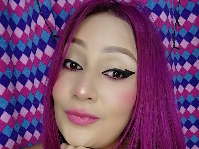 A AnyelSexy tem cabelo rosa, delineador preto, batom rosa e um sorriso adorável. Ela está segurando o queixo com a mão. Seus olhos estão olhando para a câmera..