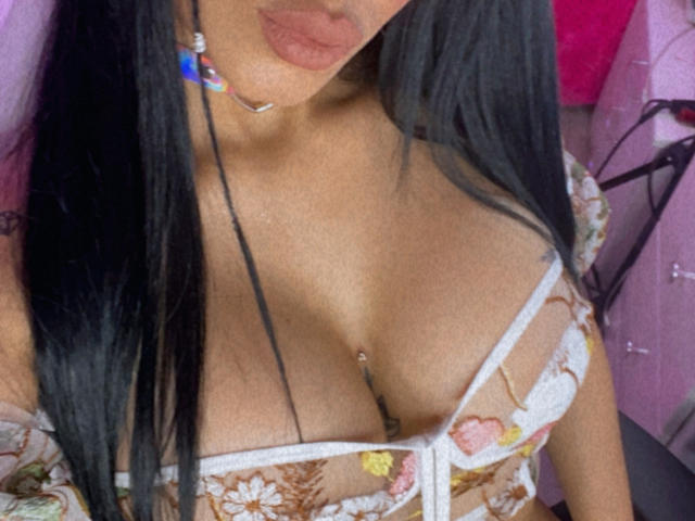OnniOconor's tits in a sheer floral bra, with a belly button piercing and black hair, wearing a colorful collar, showing plump pink lips and a tattoo on the right.