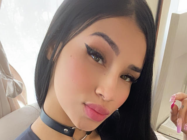 OnniOconor has black hair, green eyes, and pink lips, wearing a black choker with silver studs, and has bold, winged eyeliner. A hand with pink nails is on the right.