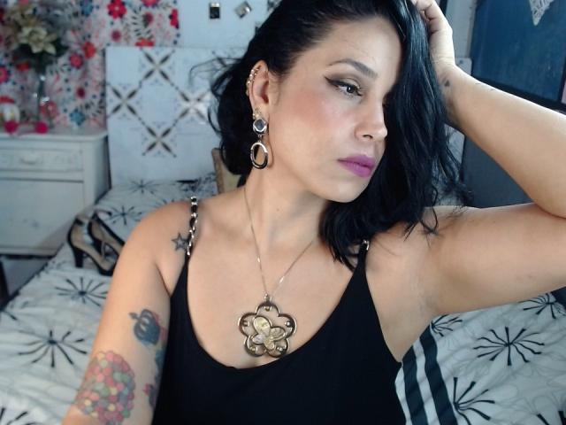 MaturesSugar69 com cabelo preto, batom rosa e regata preta, mostrando tatuagens, brincos e um colar grande com flores, com a mão no cabelo, olhando para o lado. Seios visíveis. Cenário de quarto..
