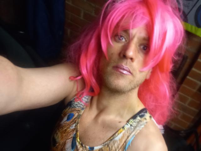 SissyMichelle, con una peluca rosa brillante, lápiz labial morado y una blusa sin mangas con estampado, tomando un selfie. Se ve un brazo, el cabello está ligeramente despeinado y tiene una expresión facial neutra..