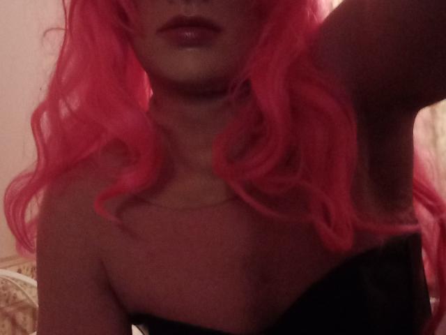 SissyMichelle, con cabello rizado rosa, labios rosados brillantes y un top negro sin tirantes, mostrando un brazo levantado, con el hueso del cuello y la parte superior del pecho visibles..