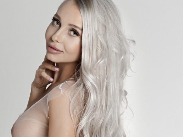 SexyBianka with long, wavy, silver hair, light skin, and green eyes, looking over her shoulder, wearing a sheer top, touching her chin, smiling softly.