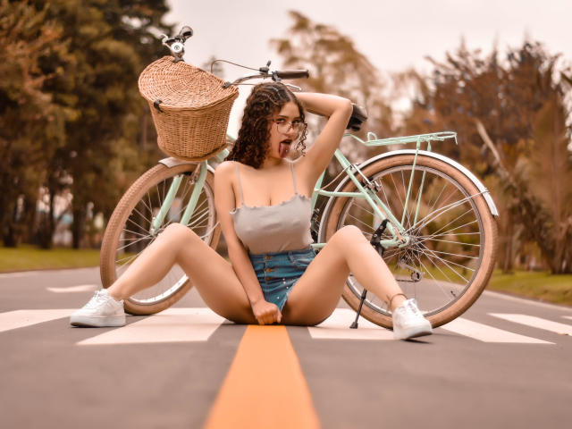 ValeryScot sitting on a road with a green bicycle, legs spread, tongue out, wearing a gray tank top, denim shorts, and white sneakers, with a wicker basket on the bike.