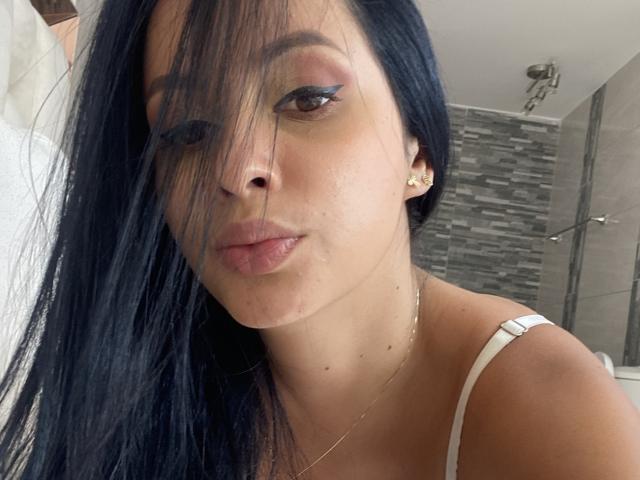 EmilyParkerr has long black hair covering one eye, light pink lips, and a nose piercing. She wears a white strap top, gold earrings, and a delicate necklace.