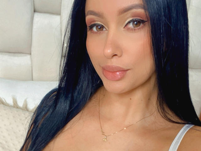 EmilyParkerr has long black hair, brown eyes, and glossy pink lips. She's wearing a thin gold necklace and a white tank top, showing her small tits.