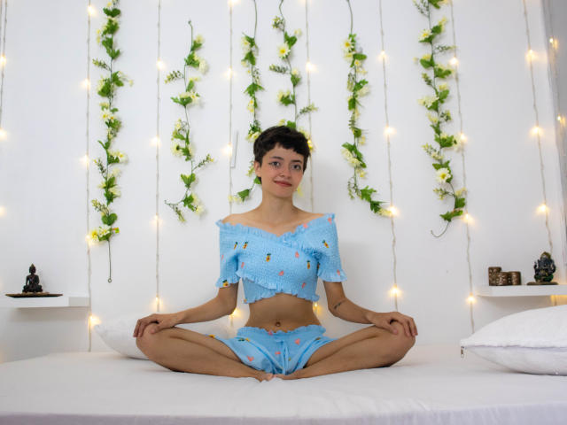 LadyRiozi sitting cross-legged on a bed, wearing a blue off-shoulder crop top and matching shorts, with small tits and a cute smile, surrounded by fairy lights and green vines.
