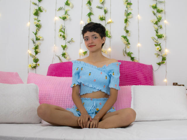 LadyRiozi está sentada con las piernas cruzadas en una cama, vestida con una camiseta corta azul con un hombro descubierto y pantalones cortos a juego, con almohadas rosas y blancas, con el pelo corto y oscuro, y sonriendo suavemente..