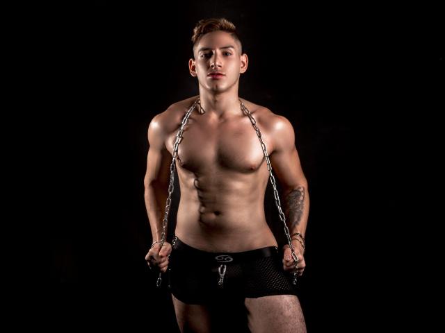 Jonathan Jeremiah, musclé, torse nu, avec des chaînes qui pendent de son cou, portant un short en maille noire, tatouage sur le bras droit, debout devant un fond sombre, cheveux bruns courts, expression sérieuse..