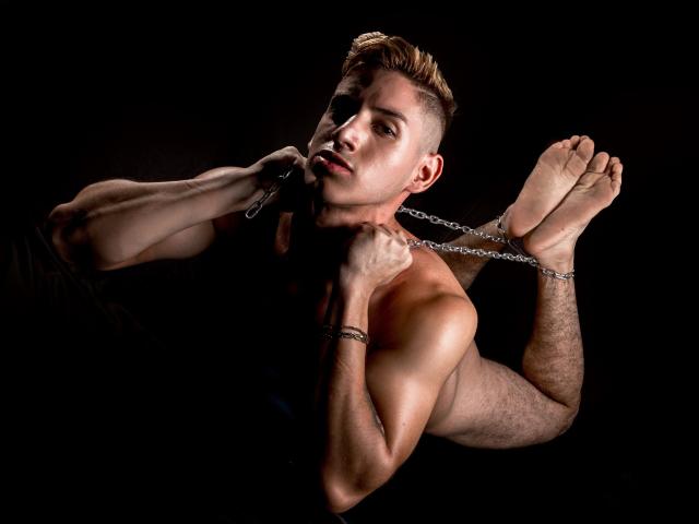JonathanJeremiah, shirtless with short blonde hair, holds a chain around his neck and wrists, his muscular arms and chest illuminated against a dark background.