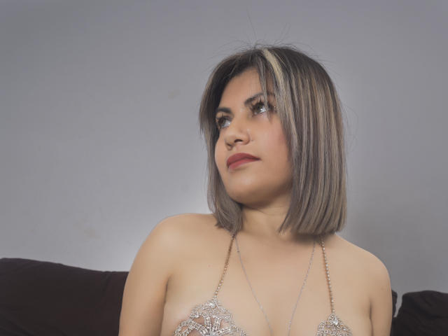 OrianaSaenz a les cheveux blonds mi-longs avec des racines foncées, elle porte un haut à encolure dégagée scintillant, elle regarde vers le haut avec un léger sourire, montrant sa petite poitrine et sa peau claire..