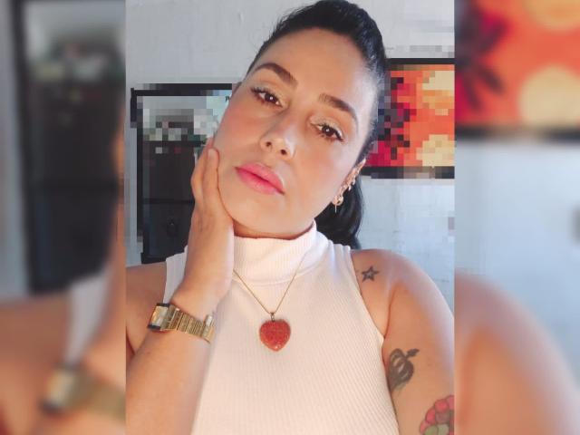 MaturesSugar69 con cabello oscuro recogido en una coleta, lápiz labial rosa, top blanco sin mangas, collar con forma de corazón rojo, reloj dorado, tatuajes en el brazo, mano en la mejilla, expresión seria, reflejo en el espejo..