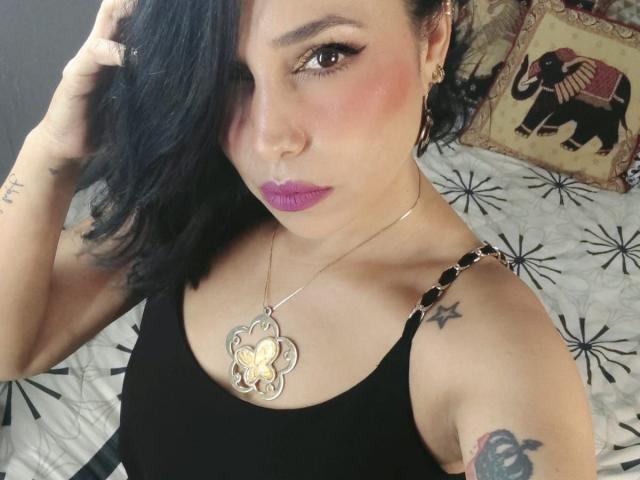 MaturesSugar69 com cabelo preto, batom roxo, camiseta de alça preta, colar com borboleta, tatuagens e brincos, olhando para a câmera, com a mão no cabelo, na cama com lençóis estampados em branco e preto..