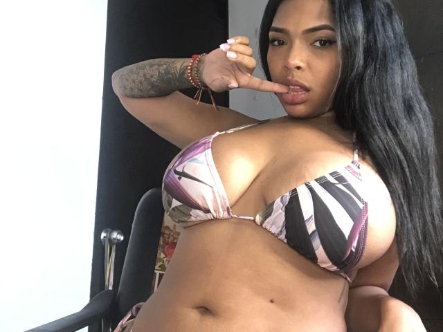 VickyGreys mit großen Brüsten in einem pink-schwarz gestreiften Bikini, mit langen schwarzen Haaren, tätowiertem Arm und Finger im Mund, sitzend auf einem Stuhl..