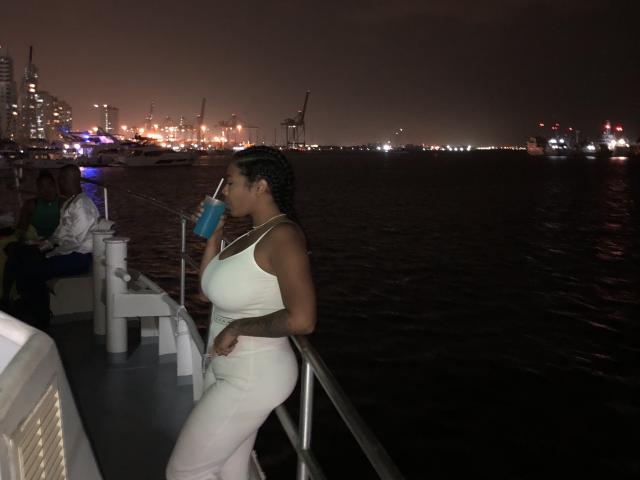 VickyGreys with dark skin, braided hair, large tits in a white tank top and pants, drinking a blue drink on a boat at night, holding rail with right hand.