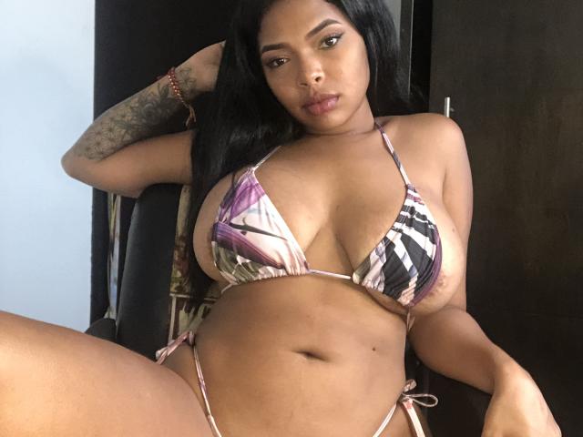VickyGreys with big boobs, wearing a colorful, patterned bikini, sitting with one arm behind her head, black hair, tattoo on her arm, looking at the camera, in a room.