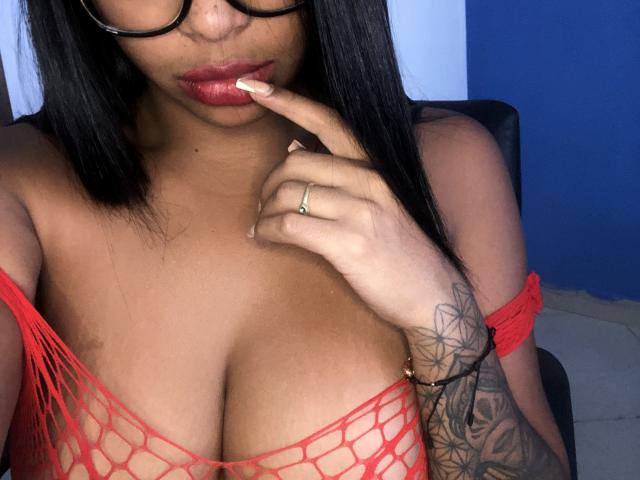 VickyGreys com pele escura e blusa de rede vermelha, mostrando seios grandes, cabelo preto, óculos, dedo perto dos lábios vermelhos, tatuagem no braço direito, anel no dedo, sentada em uma cadeira..