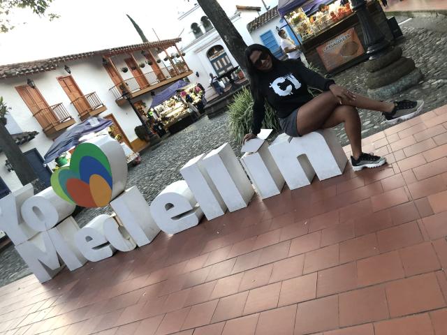 VickyGreys with dark skin, black shirt, and shorts, sitting on white "Melimn" letters, holding paper, wearing black and white sneakers, in a cobblestone and brick area.