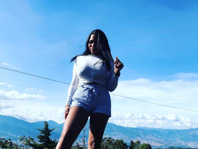 VickyGreys has long black hair, white long-sleeve shirt, and gray shorts, standing outdoors, showing off her ass and tits, with a blue sky and mountains in the background.