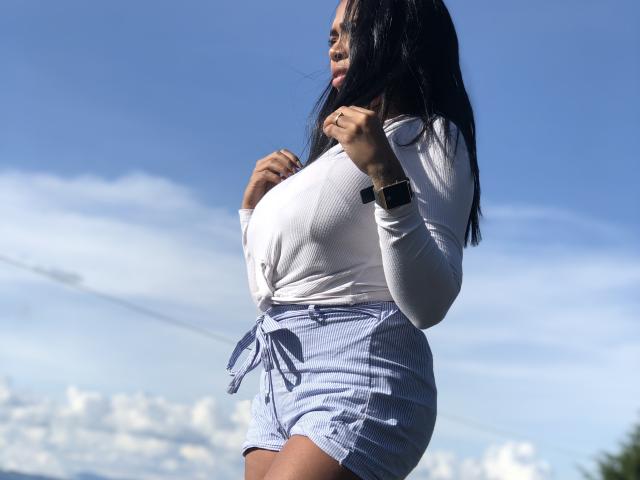 VickyGreys has big boobs, wearing a white long-sleeve top and light blue shorts, with long black hair, and a smartwatch on her right wrist, standing outdoors.