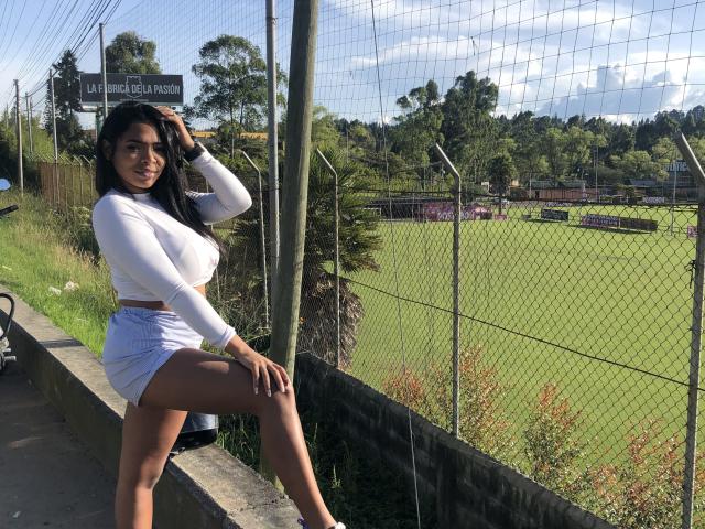 VickyGreys with large tits, wearing a white long-sleeve crop top and light blue shorts, standing by a fence, hand in hair, green field and trees in the background.