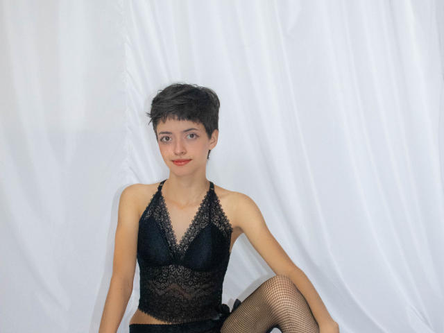LadyRiozi, avec ses courts cheveux noirs, portant un soutien-gorge en dentelle noire et des bas résille, assise contre un rideau blanc, montrant ses petites seins et un sourire discret..