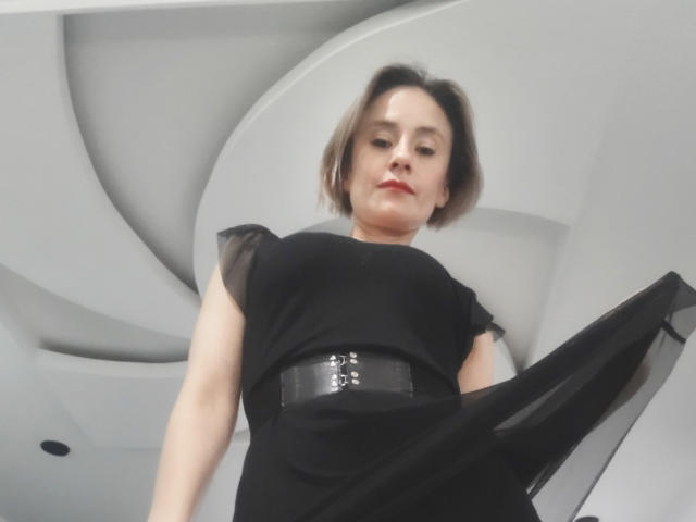 SilvyGrecor with short brown hair, black dress, and wide black belt, looking down, upshot, slight smirk, red lipstick, white ceiling with curved panels in background.