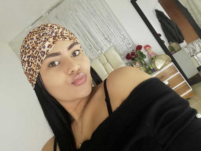 IssaHabib with leopard print headscarf, black off-shoulder top, showing bare shoulders, black hair, full lips, brown eyes, necklace, and subtle smile, in a well-lit room.