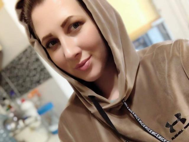 Katerina, con una capucha marrón puesta, vistiendo una camiseta Under Armour, sonriendo suavemente, con ojos marrones, piel clara, maquillaje discreto, cabello castaño parcialmente visible, en un selfie ligeramente borroso..