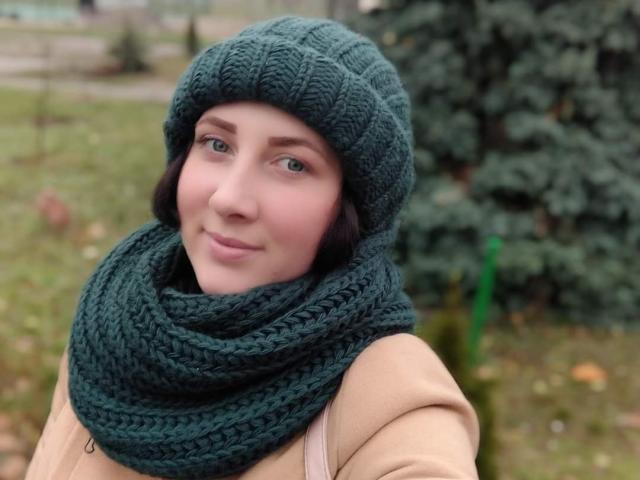 Katerina, portant un chapeau vert en tricot et une écharpe assortie, un manteau marron clair, et des cheveux courts et foncés, souriant légèrement, avec des yeux bleus et une peau claire, en extérieur, sous une lumière douce, avec une expression calme..
