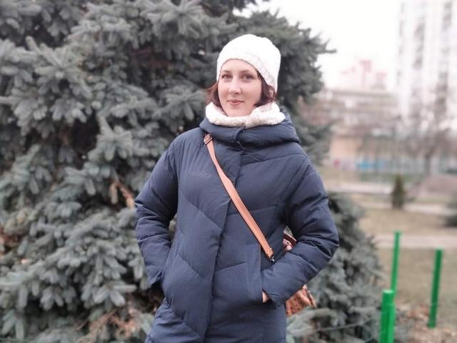 Katerina, con le mani infilate nel suo cappotto blu imbottito, un cappello bianco di lana e una borsa a tracolla marrone, è in piedi di fronte a un pino blu-verde. Fiocchi di neve visibili nell'aria..