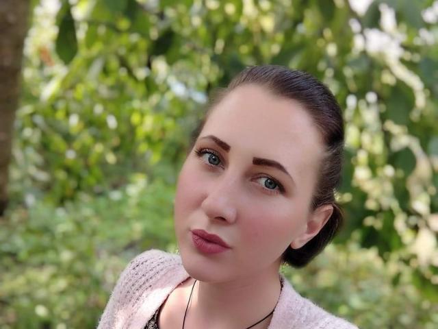 Katerina Pits tem olhos verde-claros, batom rosa e cabelo castanho preso em um coque, vestindo um suéter rosa de tricô, olhando ligeiramente para o lado com uma expressão neutra..