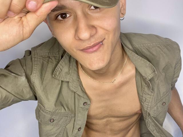 LoganD ha un corpo snello, senza camicia, con la pelle abbronzata e le costole visibili, indossa una giacca verde oliva e una catena d'oro. Indossa un berretto beige, sorride e ha un piercing al naso..
