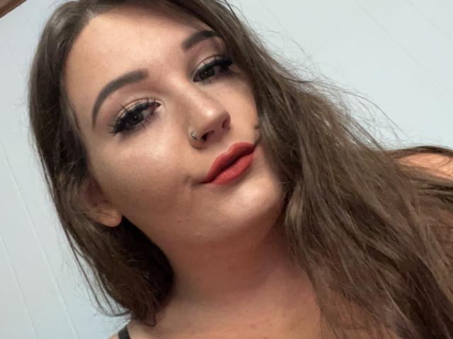 FuzyLilPeach tem cabelo castanho, um piercing no nariz e batom vermelho. Seus olhos estão semicerrados, com longos cílios pretos. Ela está fazendo uma expressão levemente emburrada, com os lábios entreabertos..