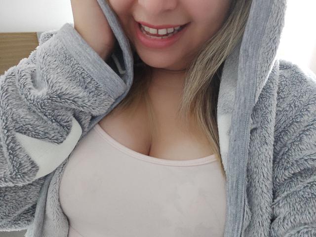 Myalewwis mit grauer, flauschiger Kapuzenjacke, rosa Tanktop, die ihre Brüste zeigt, lächelnd, sich die Kinnpartie haltend, blondes Haar, rosa Lippenstift, weiches Licht, ungezwungenes Selfie..