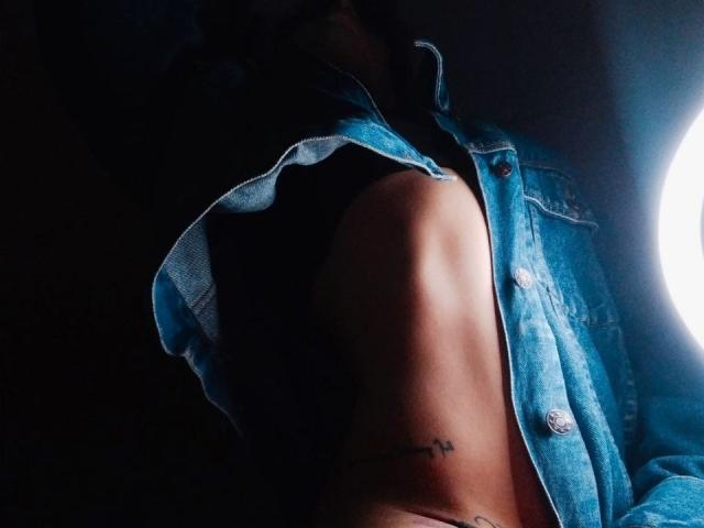 YeiSela trägt ein blaues Jeanshemd, ein schwarzes Top ohne Ärmel, hat eine freiliegende Seite, ein Tattoo am Oberarm, schwaches Licht und eine helle, runde Lichtquelle auf der rechten Seite..