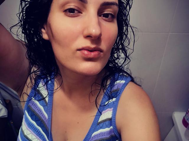 AddictedToUTS has wet, dark curly hair, light skin, and is wearing a blue and white striped tank top, with a pouty expression on her face. Her tits are small.