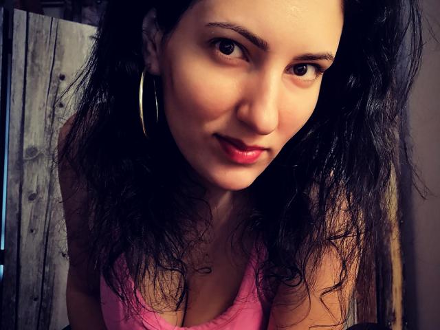 AddictedToUTS with wavy black hair, wearing pink top, large hoop earrings, and pink lipstick, looking up with a slight smile. Boobs visible, brown eyes, and light skin.