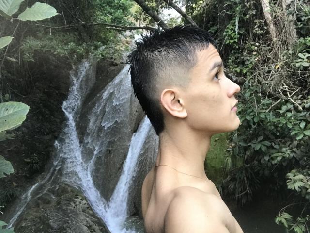 LoganD, sem camisa e com cabelo preto curto, está de lado, mostrando seu perfil e orelha. Ele está perto de uma pequena cachoeira, cercado por folhagem verde. Suas nádegas estão parcialmente visíveis..