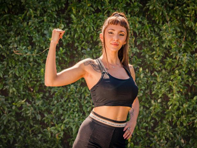 ElizaWells with a flexed bicep, wearing a black sports bra and high-waisted black pants, showing tattoos on her shoulder and upper arm, brown hair in a ponytail.
