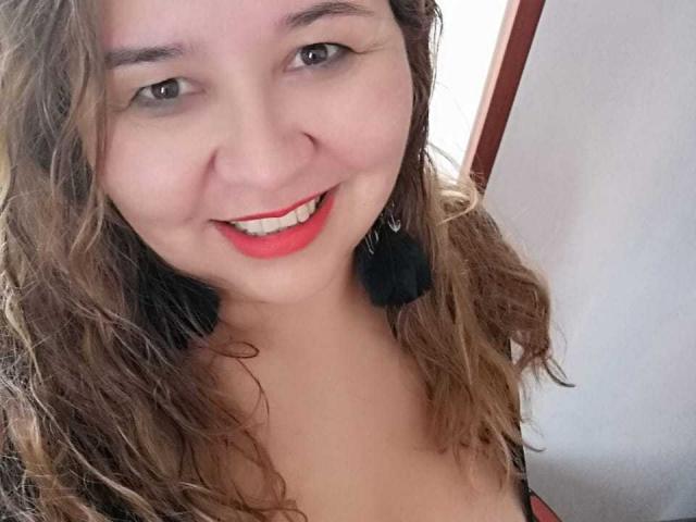 AmbarSimmons with wavy brown hair, red lipstick, and a smile, showing her tits in a selfie. She's wearing black feather earrings and a red top.