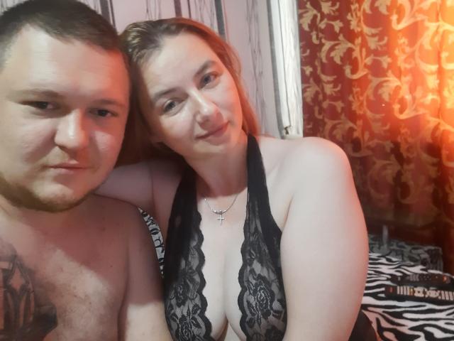 PatriciaNMike e Mike, ambos com pele clara, os seios grandes de PatriciaNMike em renda preta, Mike sem camisa com tatuagem, braço ao redor de PatriciaNMike, luz laranja, cama com padrão de zebra..