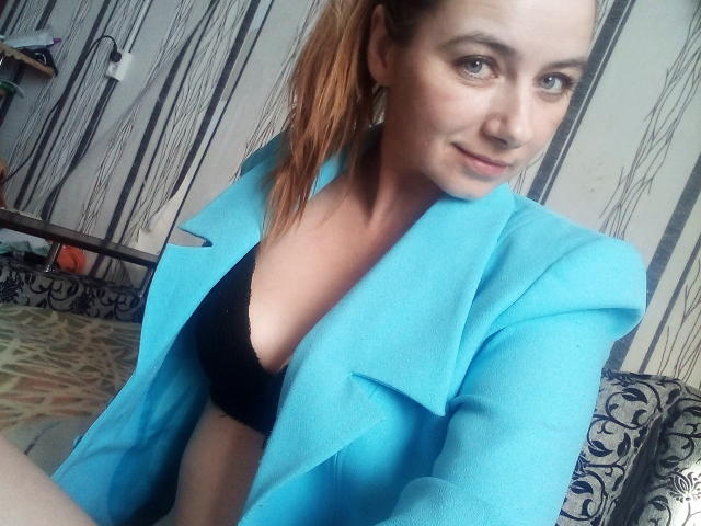 PatriciaNMike met lichtbruin haar, gekleed in een blauw blazer en een zwarte bh, waarbij haar kleine borsten zichtbaar zijn, lachend en recht in de camera kijkend. Transparante huid, lichte make-up, nonchalante pose..