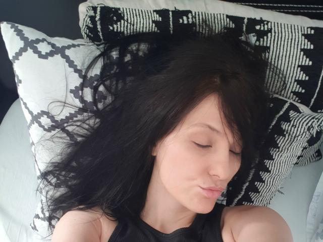JoliDanielle with closed eyes, black hair, and light skin, lying on a black and white patterned pillow, wearing a black sleeveless top, showing her nose and lips.
