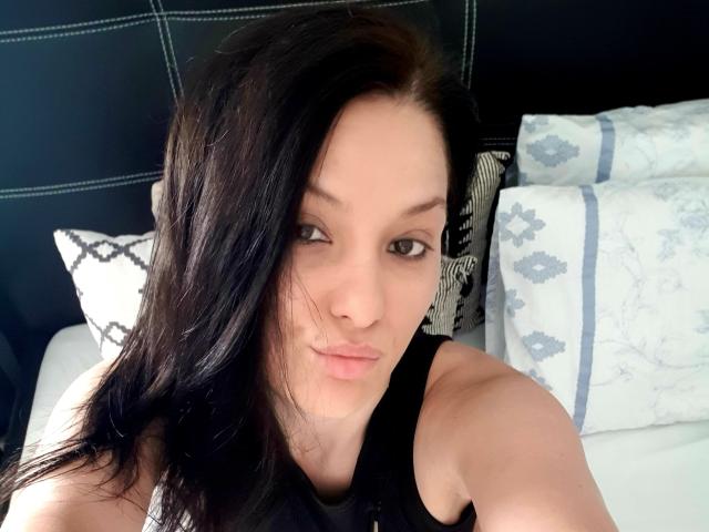JoliDanielle with black hair, pouty lips, and green eyes, wearing a black tank top, taking a selfie, showing her bare shoulders and a subtle, sexy smile.