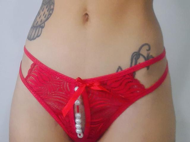 La lingerie rossa in pizzo di LolyFunX, con un filo di perle nell'area inguinale, mostra i suoi tatuaggi sul lato destro del suo ventre piatto e della parte inferiore dell'addome..
