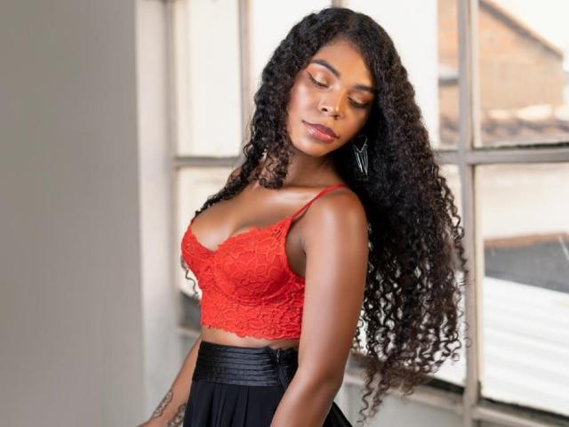 TianaParkerr, con la pelle scura, lunghi capelli ricci neri, indossa un reggiseno di pizzo rosso e una gonna nera, mostrando il seno e un tatuaggio sul braccio destro, e guarda in basso..