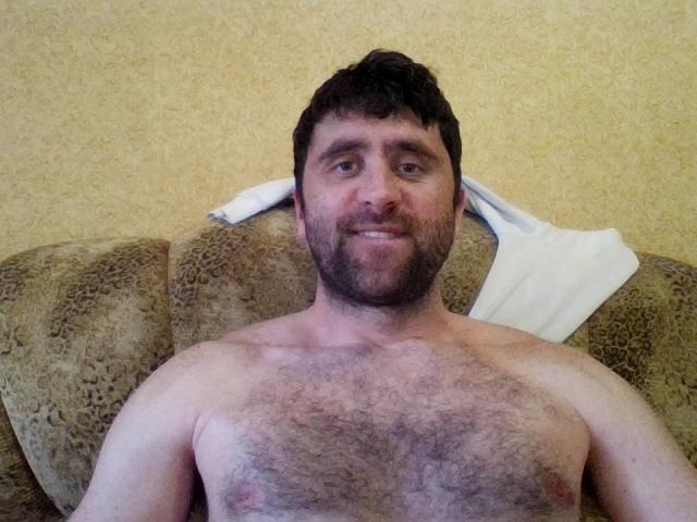 StasConnor, sem camisa, com o peito peludo, cabelo curto e escuro, e barba, sorrindo para a câmera, apoiado em um sofá estampado com uma toalha branca atrás da cabeça..